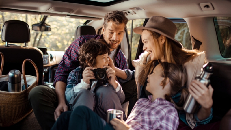 A family sits together in the back of their car, smiling and sharing drinks and a camera during a road trip